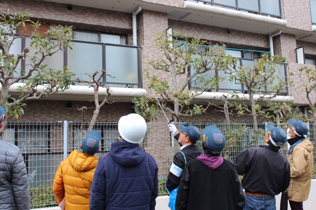 マンションの外壁を見上げ、専門家の説明を聞きながら建物の状態を確認する参加者